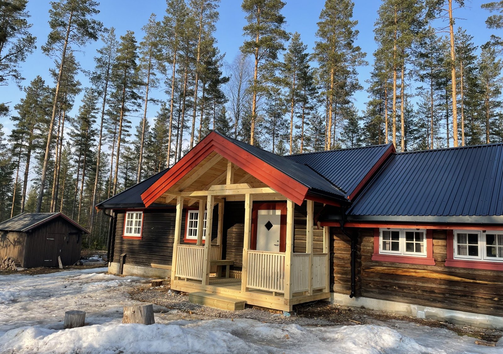 Tilbygg på tradisjonell tømmerhytte med overbygd inngangsparti i tre, omgitt av snø og furuskog.