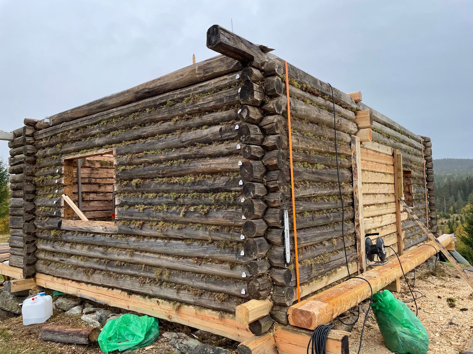 Pågående lafting av tømmerbygg med synlig mose i fugene og nummererte stokker, plassert i fjellterreng.
