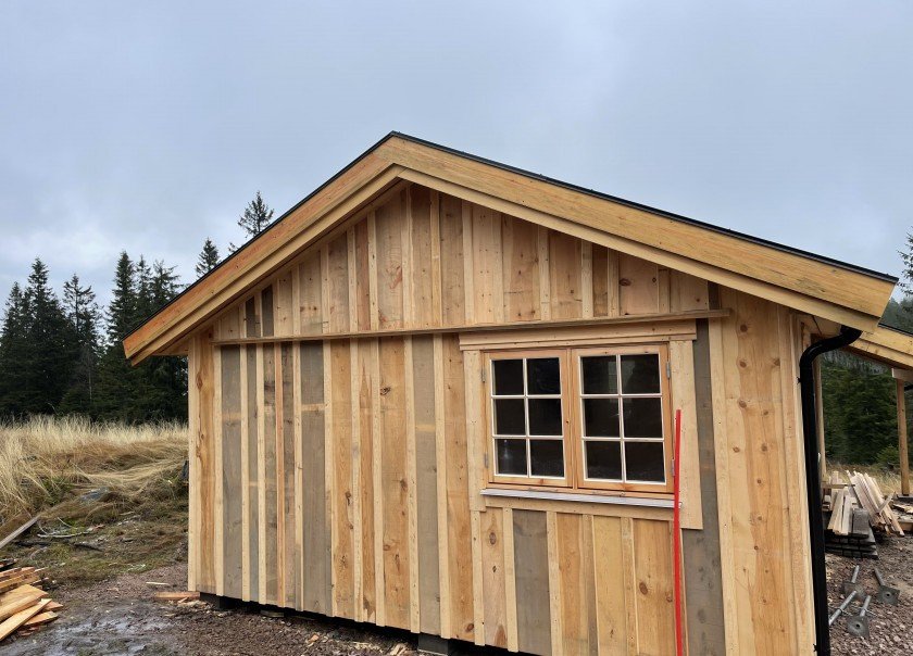 Hytte under oppføring med stående kledning i ubehandlet tre og sprossevindu, omgitt av skog og myrområde.