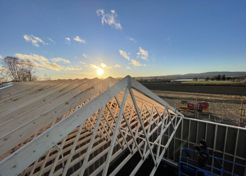 Takstoler på uthus under bygging i solnedgang, med åkerlandskap og blå himmel i bakgrunnen.