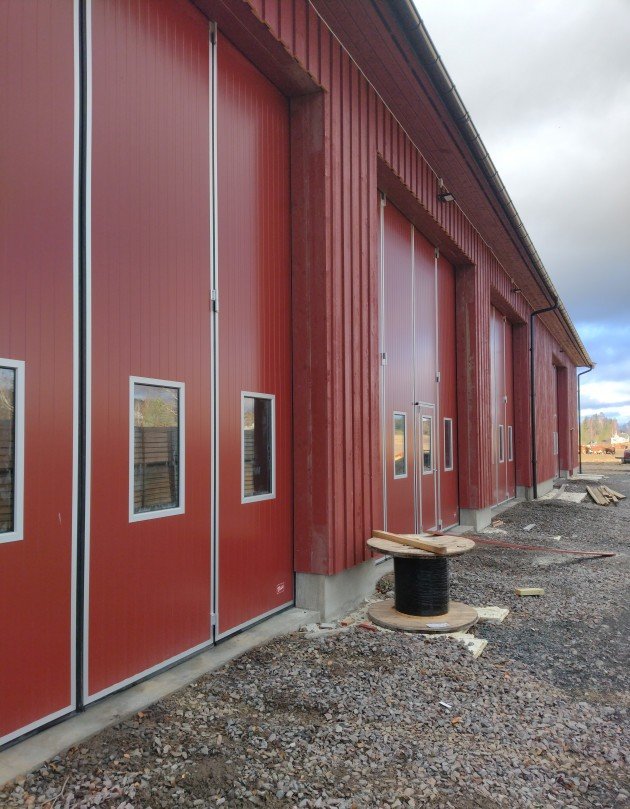 Rødmalt uthus med store porter og vinduer, stående kledning og gruslagt område foran bygget.